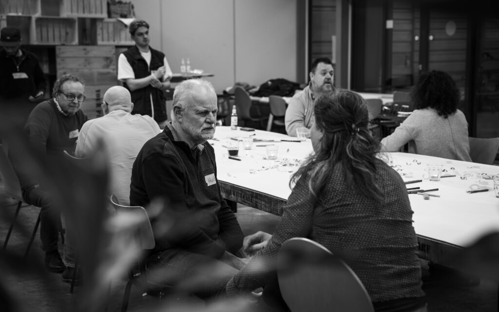 Deelnemer Huib luistert aandachtig naar Karin haar verhaal. Op de achtergrond zijn de overige deelnemers één op één met elkaar in gesprek.