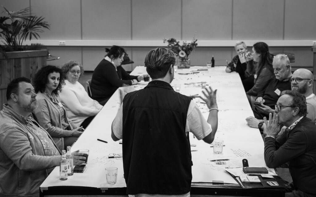 Alle deelnemers aan tafel die kijken en luisteren naar organisator Luke op de voorgrond. Hij vertelt de volgende opdracht.