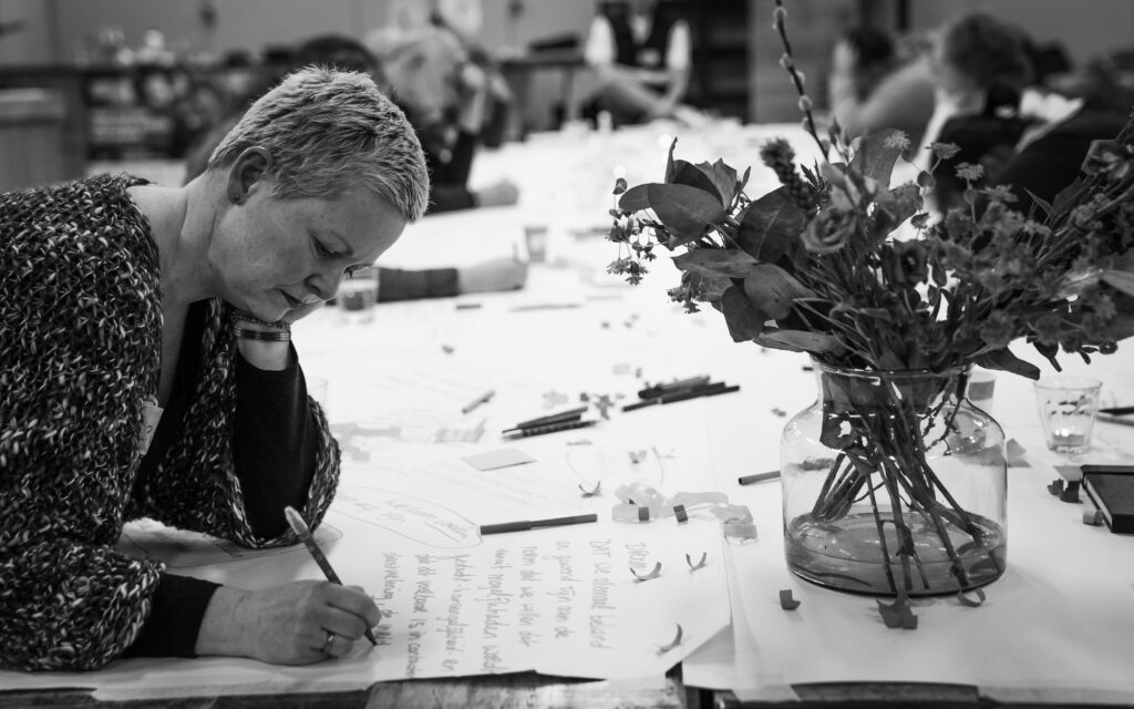Deelnemer Desireé is flink aan het schrijven op haar vel papier. Tegenover haar staat een bos met bloemen op tafel.