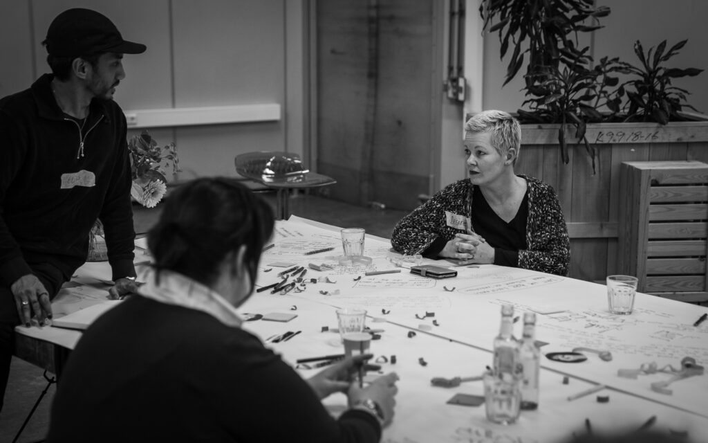Deelnemer Desireé zit aan tafel en bespreekt haar verhaal met organisator Elias, die op tafel zit. Elias kijkt terug naar Desireé.